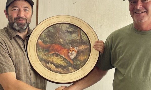 Zwei Männer halten gemeinsam ein gerahmtes Rundbild eines Fuchses in natürlicher Umgebung. Sie schütteln sich die Hand vor einer Holzwand und lächeln in die Kamera.